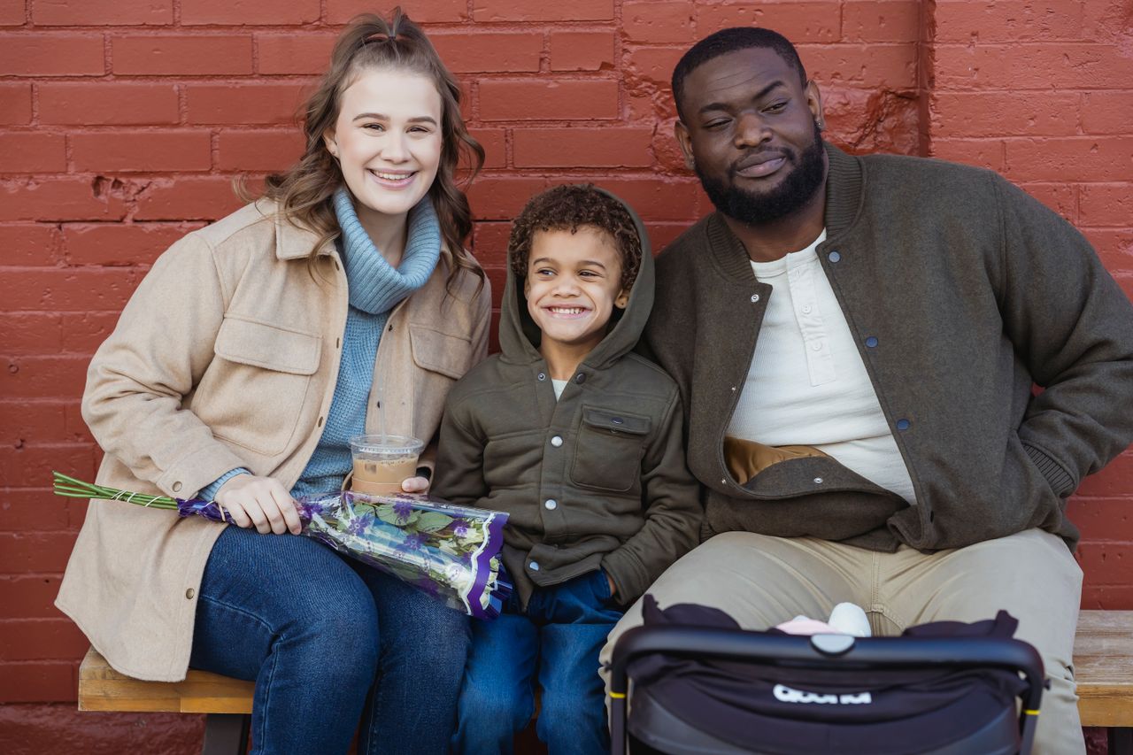 Happy multiracial couple and cute son wearing warm clothes sitting together on bench against shabby brick wall